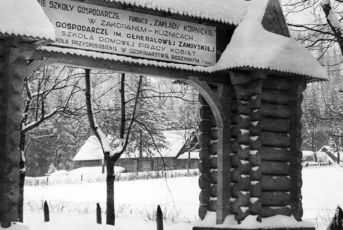 Brama Szkół Gospodarczych Fundacji Zakłady Kórnickie w Zakopanem: Architektonicznie bogata brama wykonana z drewna, pokryta śniegiem. Na górze widnieje tablica z napisem informującym o Szkołach Gospodarczych Fundacji Zakłady Kórnickie w Zakopanem. W tle widać zaśnieżony krajobraz z drzewami i domami.