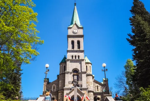 Kościół pw. Świętej Rodziny przy ul. Krupówki w Zakopanem (nowe zdjęcie): Współczesna fotografia kościoła z charakterystyczną wieżą, uchwycona w słoneczny dzień. Widok od schodów prowadzących do głównego wejścia, otoczonego drzewami i latarniami.