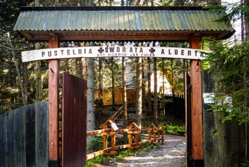 Pustelnia św. Brata Alberta na Kalatówkach: Wejście do pustelni św. Brata Alberta w Zakopanem, z charakterystycznym drewnianym łukiem nad bramą. W tle widać las i ścieżkę prowadzącą do zabudowań.