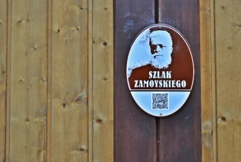 Tabliczka Szlaku Zamoyskiego: Fotografia przedstawia owalną tabliczkę na drewnianej ścianie, oznaczającą Szlak Zamoyskiego. Na tabliczce znajduje się wizerunek Władysława Zamoyskiego, napis 