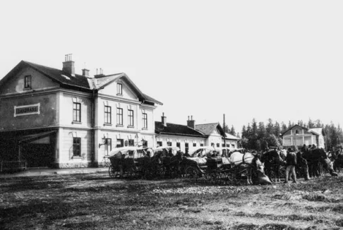 Dworzec kolejowy w Zakopanem z postojem pojazdów konnych (stare zdjęcie): Czarno-biała fotografia przedstawiająca dworzec kolejowy w Zakopanem, przed którym znajduje się postój pojazdów konnych. Widoczne są liczne wozy zaprzężone w konie oraz grupy ludzi zgromadzonych wokół. Budynek dworca ma klasyczną architekturę z charakterystycznym napisem 