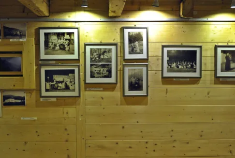 Wnętrze wystawy historycznej: Fotografia przedstawiająca ścianę wewnątrz muzeum z ekspozycją starych zdjęć w ramach. Drewniana ściana jest oświetlona, ukazując fotografie związane z historią regionu.
